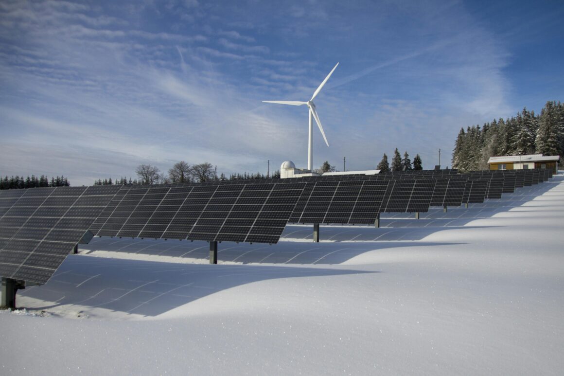 Solar panels in a snowy field with a single wind turbine behind them